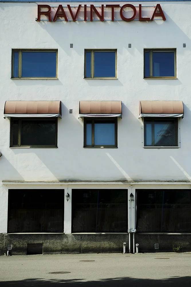 Front view of a white building with a red sign that reads "RAVINTOLA" on top, having three rows of windows, some with pink awnings, and a ground level with large black windows or doors.