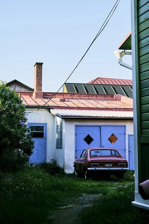 Old red car parked in front of a house with blue doors and windows, green grass and trees, and neighboring houses with red and green roofs.