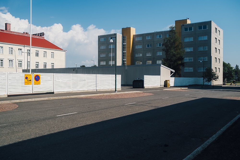An urban street scene with a white fence, a dumpster, and a multi-story apartment building under a partly cloudy sky.