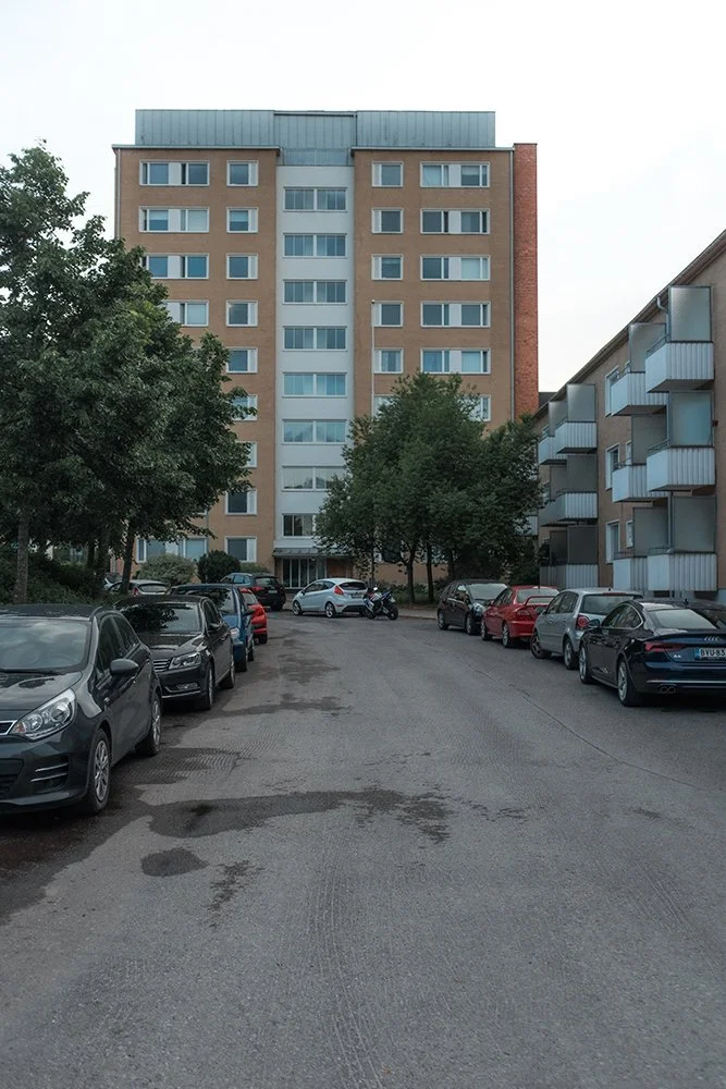 Residential apartment building with several parked cars in front and trees lining the parking area.