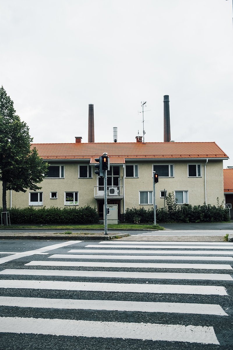 A pedestrian crosswalk in front of a residential building with a red-tiled roof and three smokestacks in the background. Traffic lights are visible.