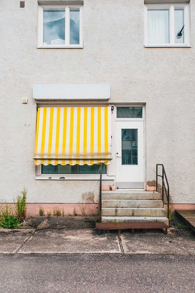 Exterior of a building with a yellow and white striped retractable awning over a window, a white door with a small window, and two steps leading up to the door