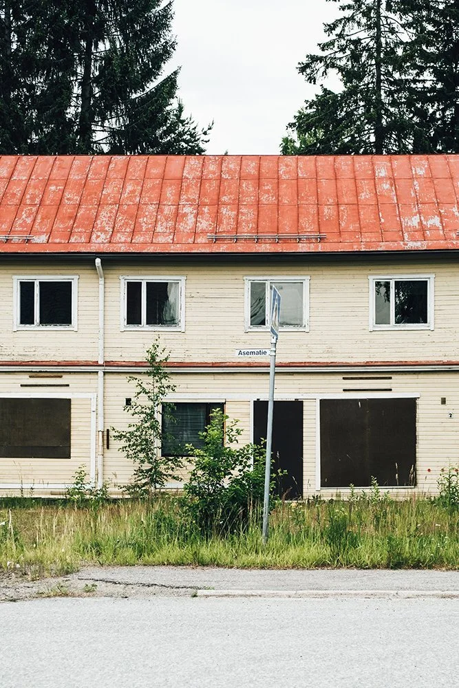 An abandoned two-story building with boarded-up windows, a red rusted metal roof, and overgrown grass in front. A street sign with partially visible text is in the foreground.