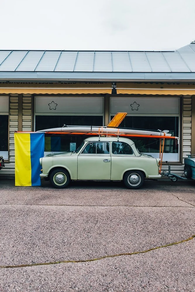 An old beige car parked in front of a house, equipped with a surfboard and a kayak on its roof. A Ukrainian flag is draped beside the car.