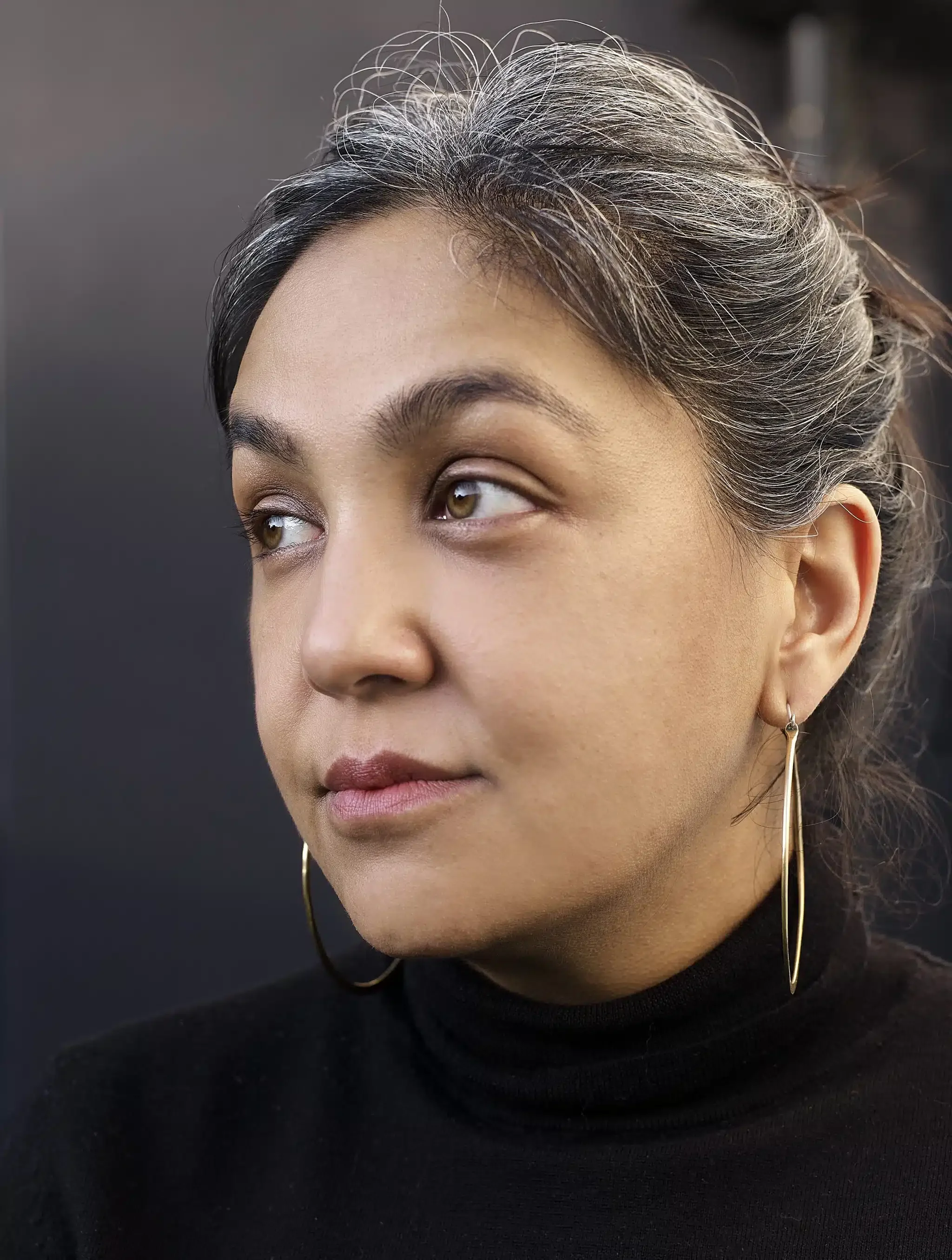 A close-up of a young Asian woman's face showing her right eye, shoulder-length black hair with streaks of gray, and a gold hoop earring on her right ear. She is wearing a black top.