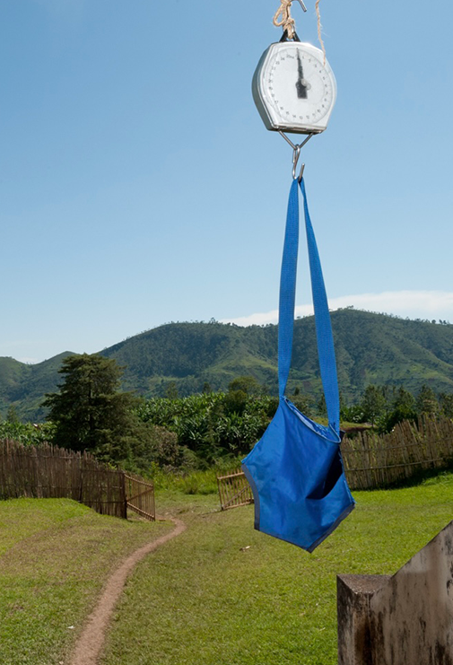 A hanging weight scales with a blue bag attached to the strap, outdoors with green hills and a dirt path in the background.