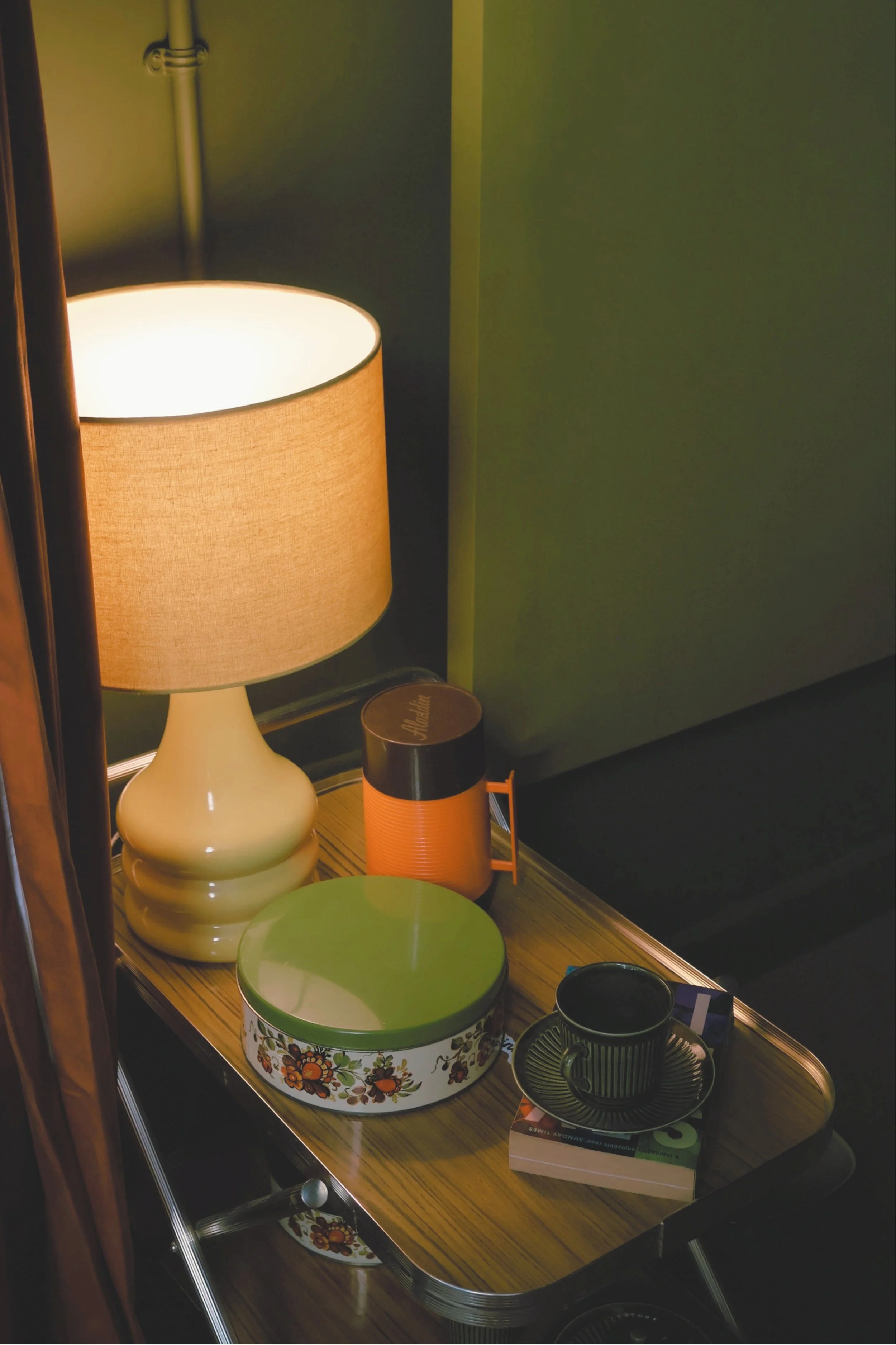 A wooden display shelf with various containers and decorative items, including a lampshade, a floral-patterned bowl, a small orange container, and a black mug, set against a green wall.