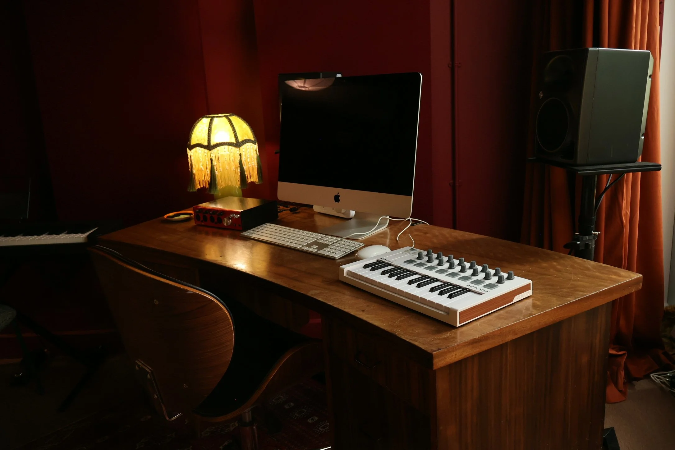 A music production workspace with a computer, MIDI keyboard, speaker, and recording equipment on a wooden desk, illuminated by a vintage yellow lamp with a fringe shade.