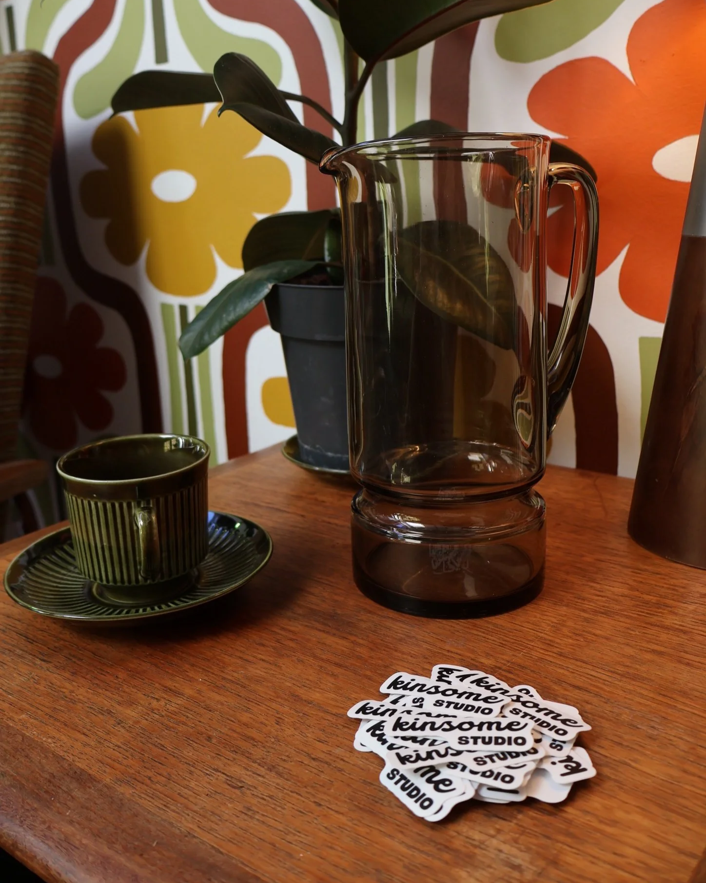 A wooden table with a green and black striped cup and saucer, a transparent glass pitcher, a small potted plant with dark green leaves, and a stack of stickers that read 'kinsome studio'. The background has a floral-patterned wallpaper with large, stylized flowers in green, yellow, orange, and white.