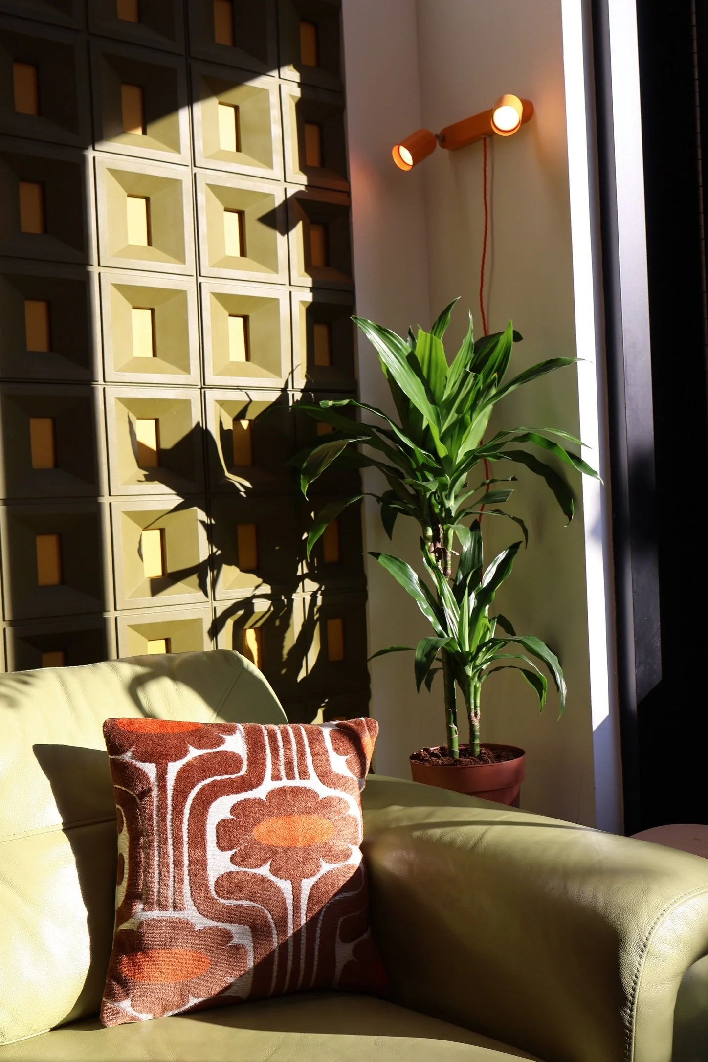 A room corner with a yellow sofa, a decorative pillow with a brown and orange pattern, a green potted plant, a wall with square geometric wooden accents, and a wall-mounted light fixture with two cylindrical wooden lights. Sunlight streams through a nearby window, casting shadows on the wall.