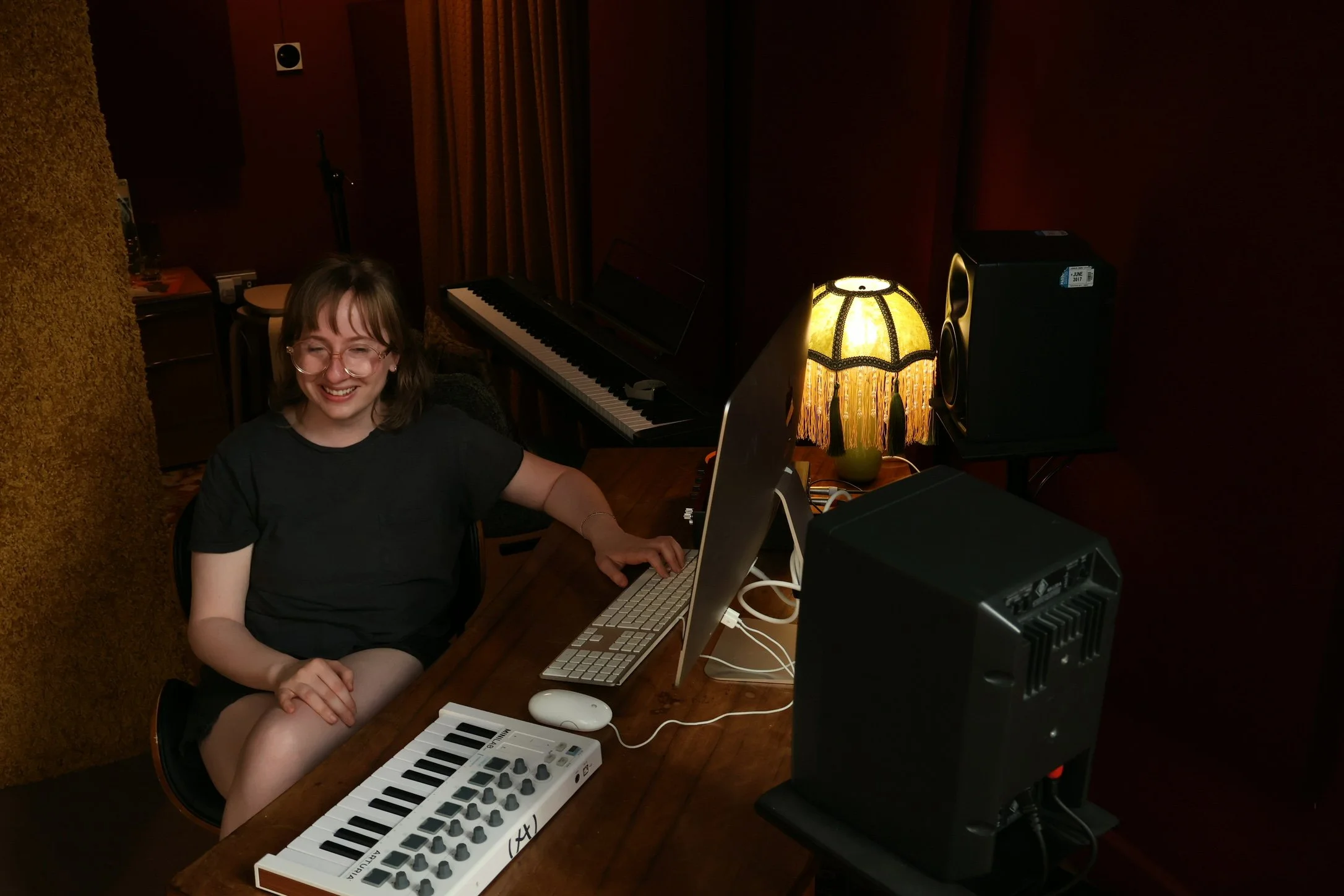 A woman with glasses smiling at a music studio surrounded by musical equipment, including a keyboard, computer, and speakers, with a vintage lamp providing warm lighting.