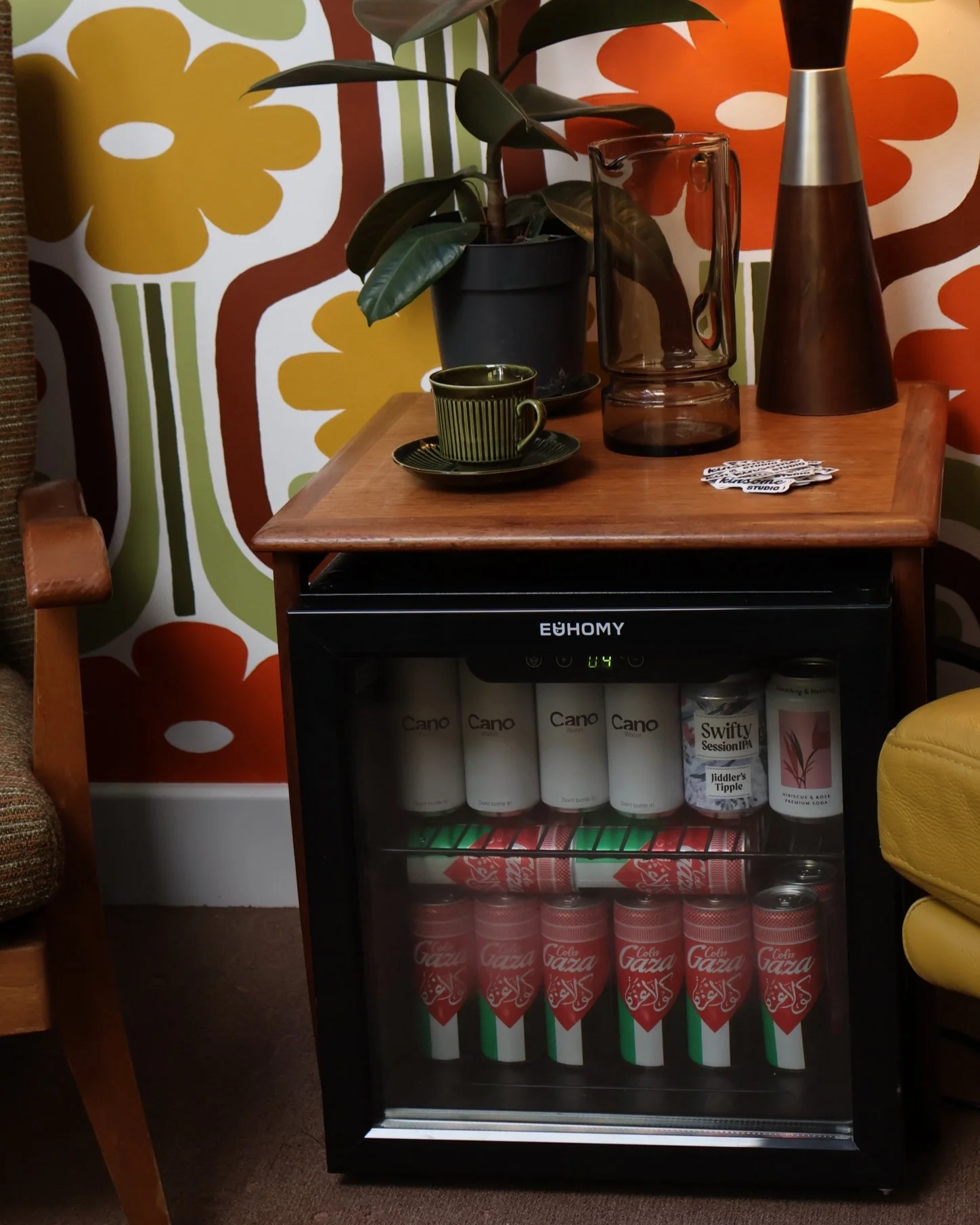 A small fridge with drinks, a potted plant, a glass pitcher, a cup and saucer, and a decorative lamp and spray bottle on top, set against a colorful floral wallpaper.