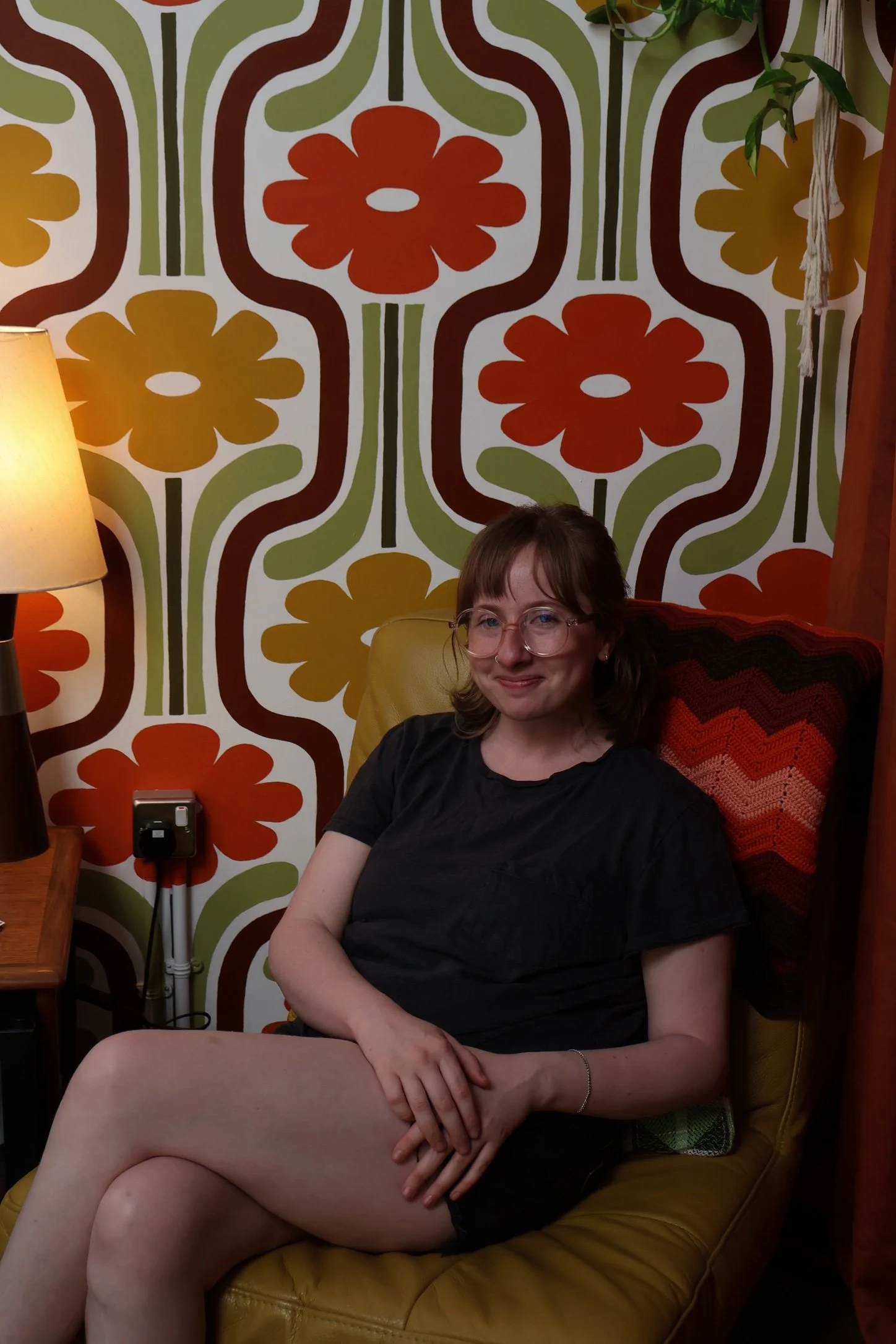A woman with glasses and blond hair sitting on a yellow leather chair in a room with a retro floral wallpaper background. She is wearing a black top, smiling, with her legs crossed, one arm resting on her lap, and the other on the chair.
