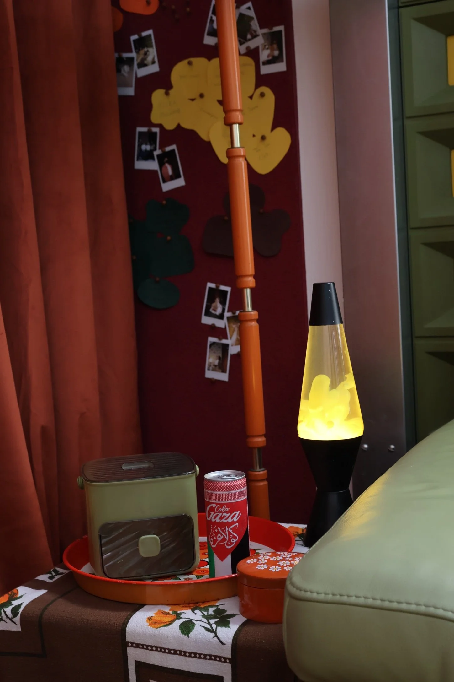 A cluttered living space with a yellow lava lamp, a can of Cola Gaza soda, a red plastic tray, and various other items on a table with a floral tablecloth. The background shows a wall with photos, a colorful lamp, and a tall orange pole.