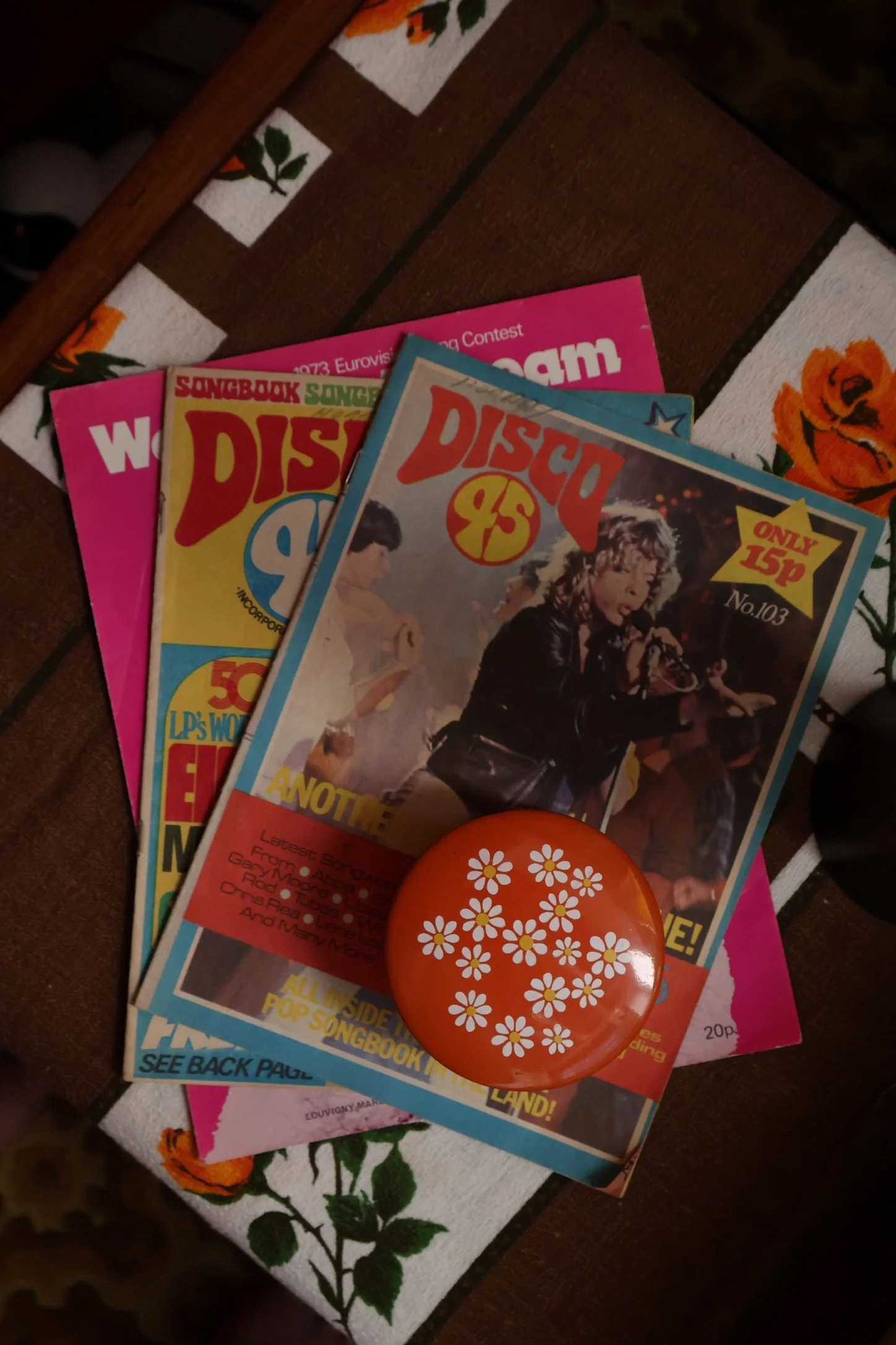 A small collection of magazines and a round orange object with white daisy pattern, placed on a wooden surface next to a floral-patterned cloth.