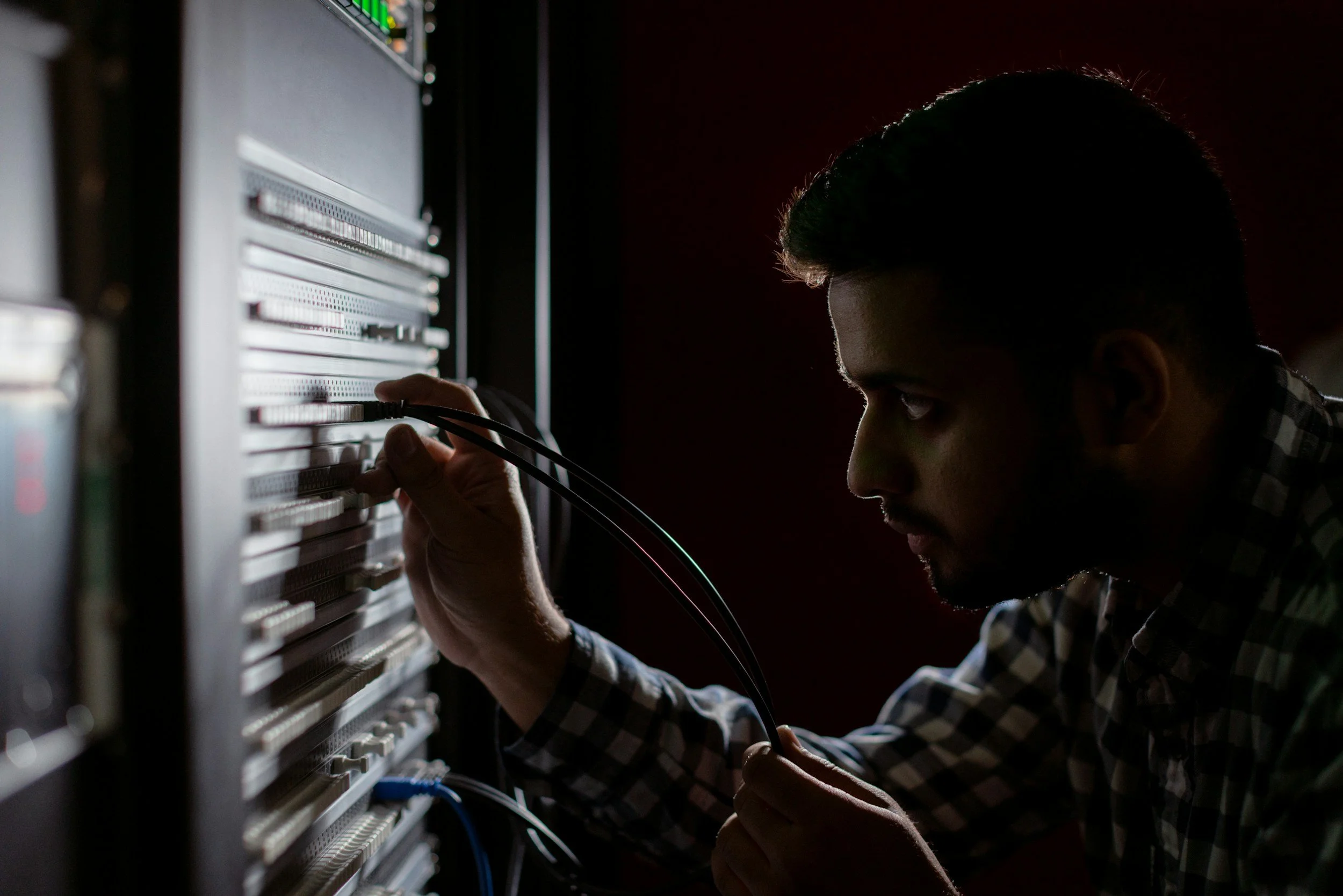 A man working on server equipment in a dark room, connecting cables to a rack of electronic devices.