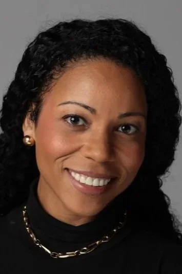 A woman with dark curly hair smiling at the camera, wearing pearl earrings, a black top, and a gold chain necklace.