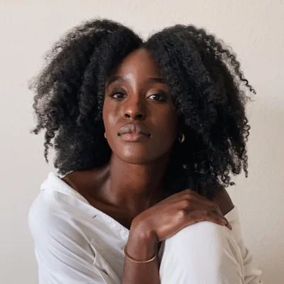 Portrait of a woman with curly black hair, wearing a white top, posing against a plain light-colored background.