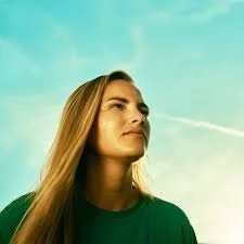 Young woman with long hair looking thoughtfully into the distance outdoors, with a blue sky background.