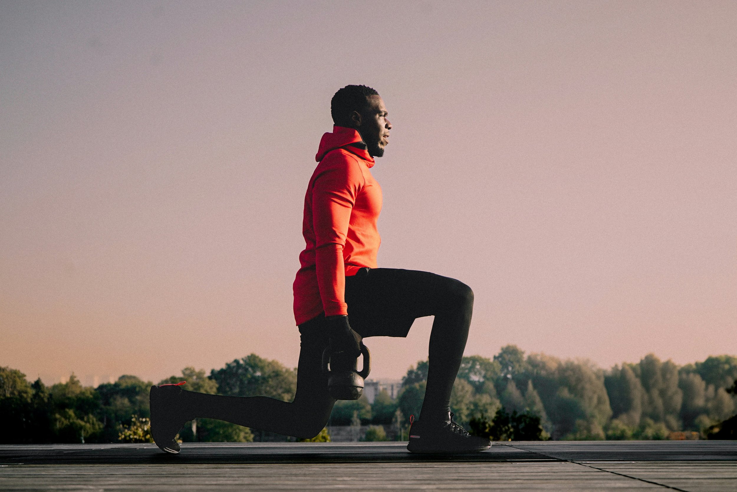 A man wearing a red hoodie and black pants performing a yoga pose on a black yoga block outdoors during sunset, with trees and a light sky in the background.