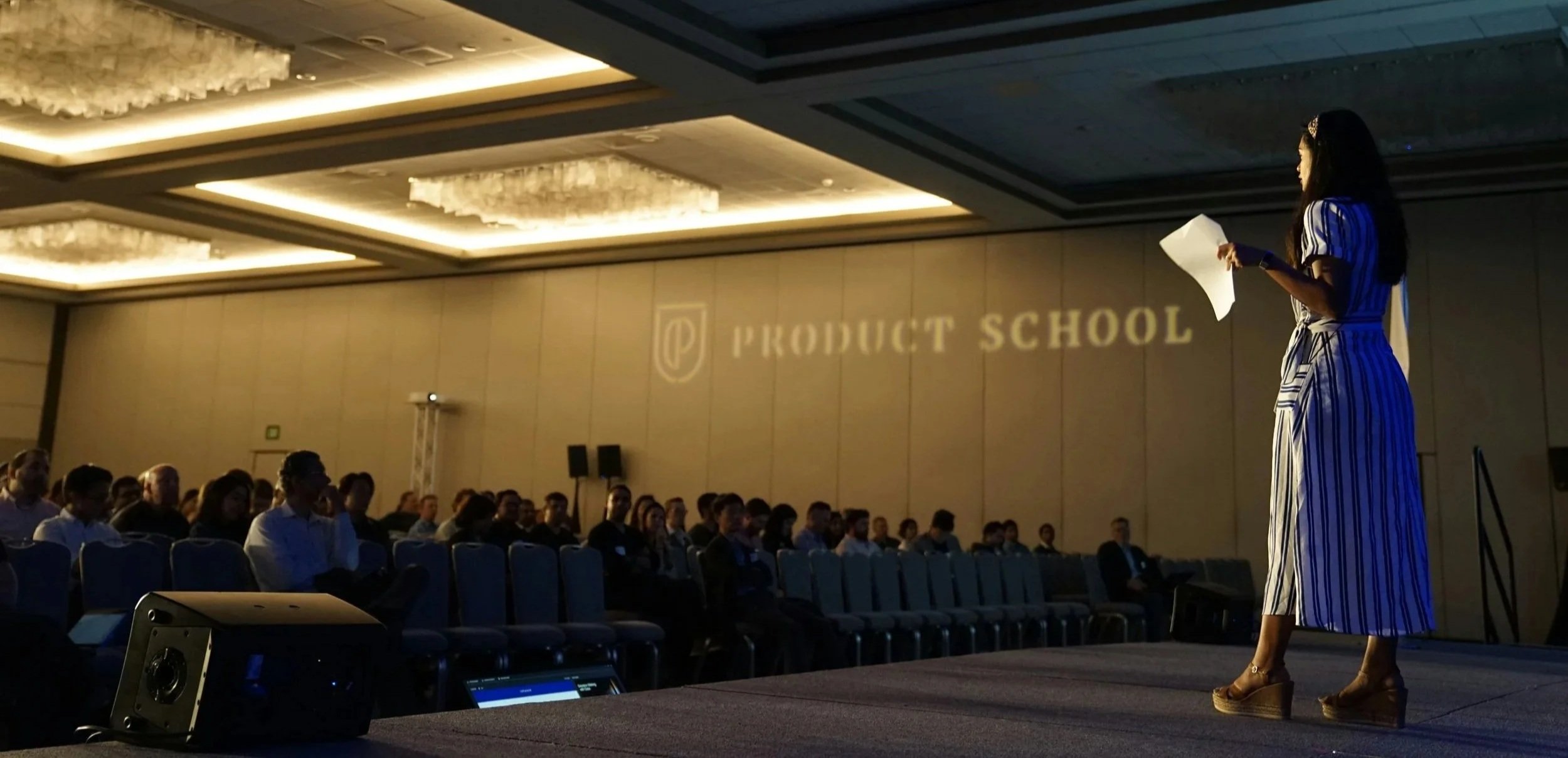 A woman in a striped dress and high heels is standing on stage holding a microphone and a piece of paper at a conference or seminar titled 'Product School,' with an audience seated at tables in front of her. The room has a large chandelier and projected text on the wall.