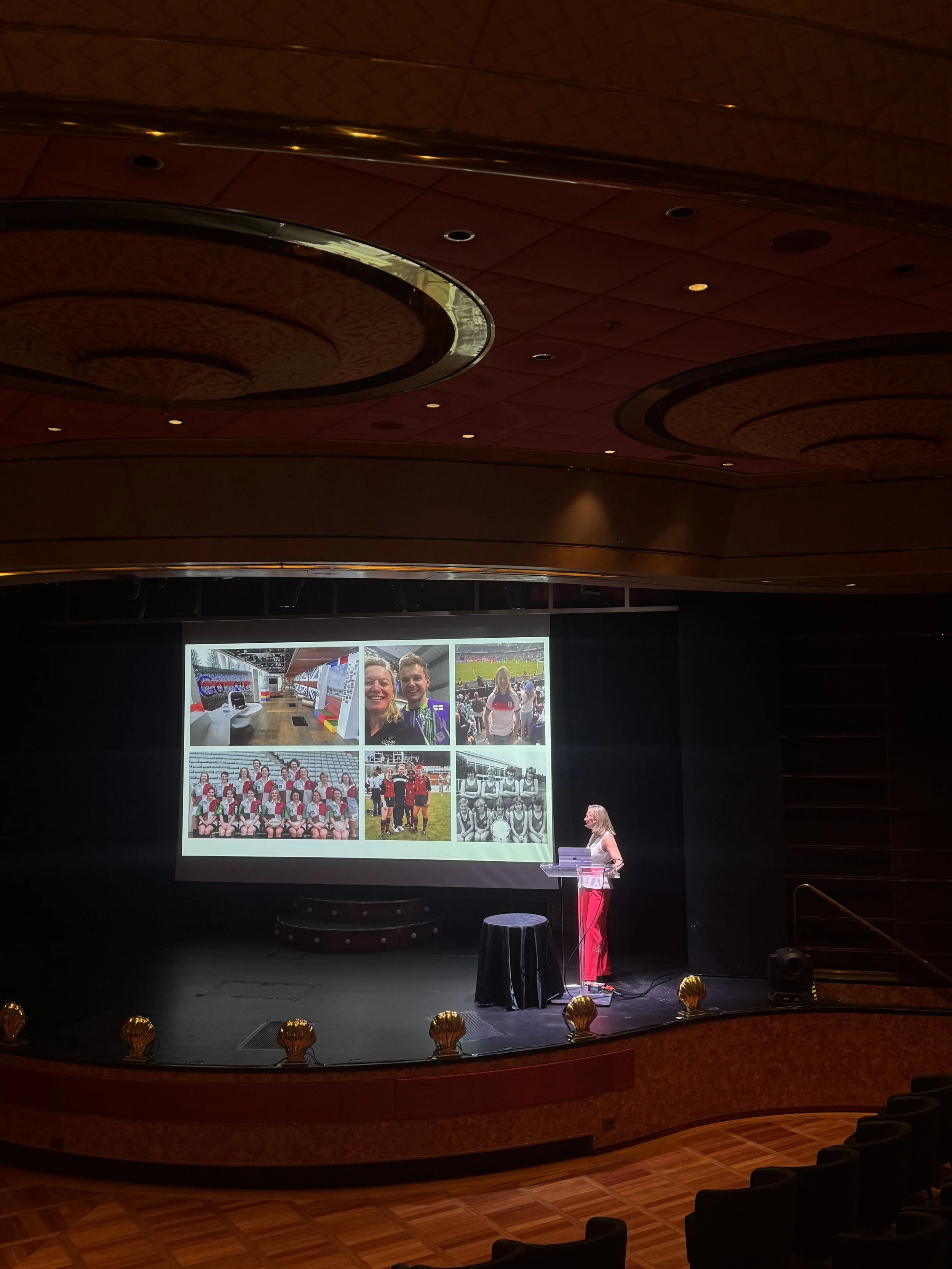 Woman giving a presentation on stage with a large screen displaying photos of people and events behind her. The auditorium is dimly lit with empty seats in the foreground.