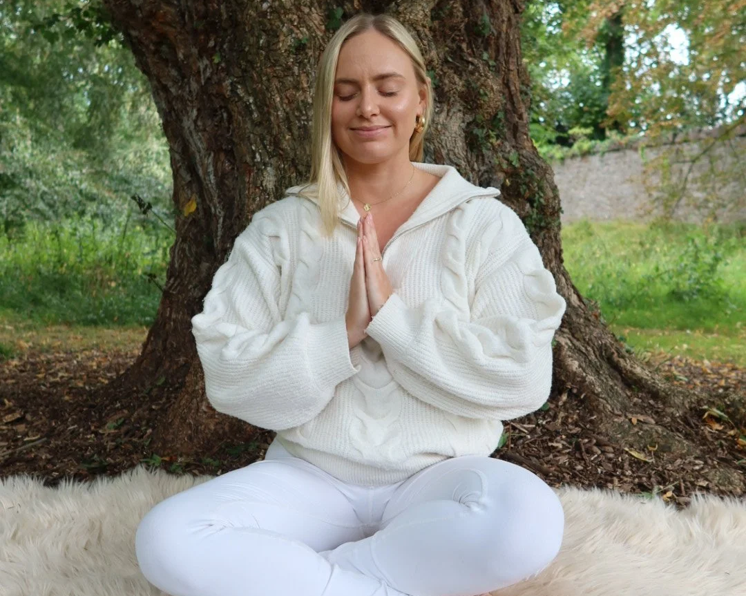 Woman meditating outdoors in a park, sitting cross-legged on a white fur rug with a large tree trunk behind her.