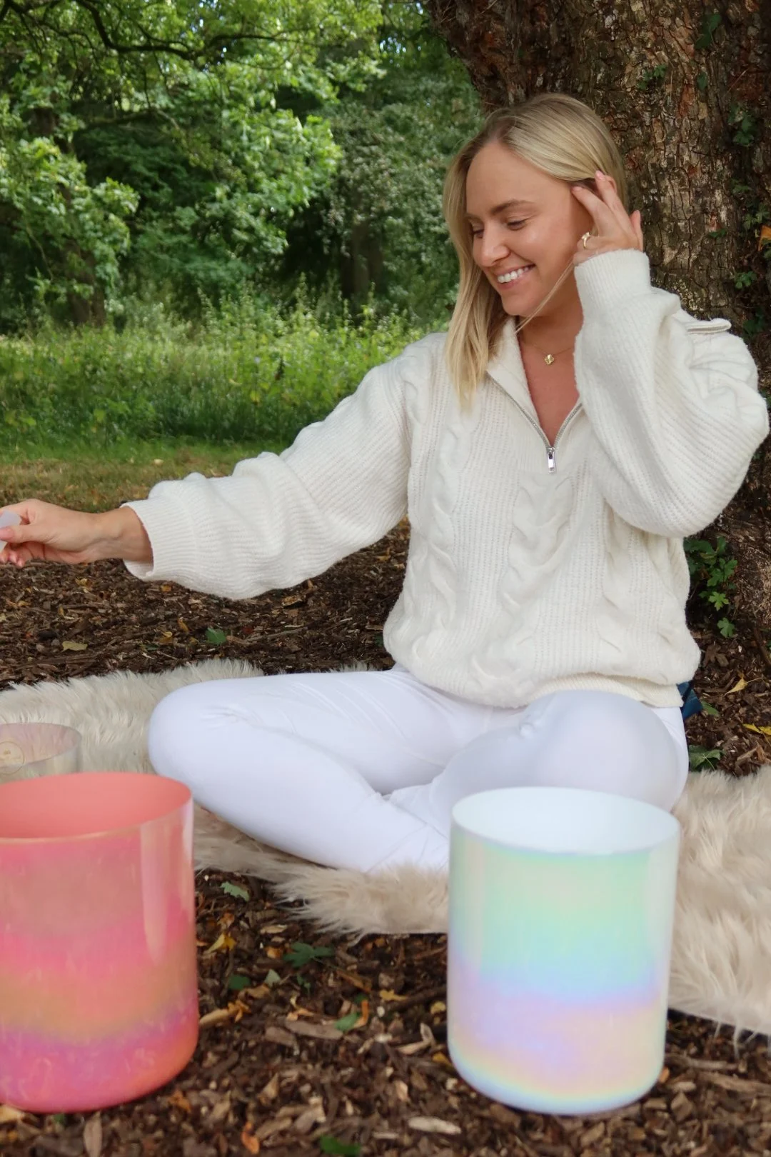 A woman sitting outdoors on a furry blanket beneath a tree, smiling. She is dressed in a white cable knit sweater and white pants, surrounded by colorful cylindrical sound bowls.