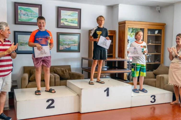 Tres niños en un podio ganadores en una ceremonia de premiación, con adultos aplaudiendo en una sala decorada con cuadros y mobiliario de madera.