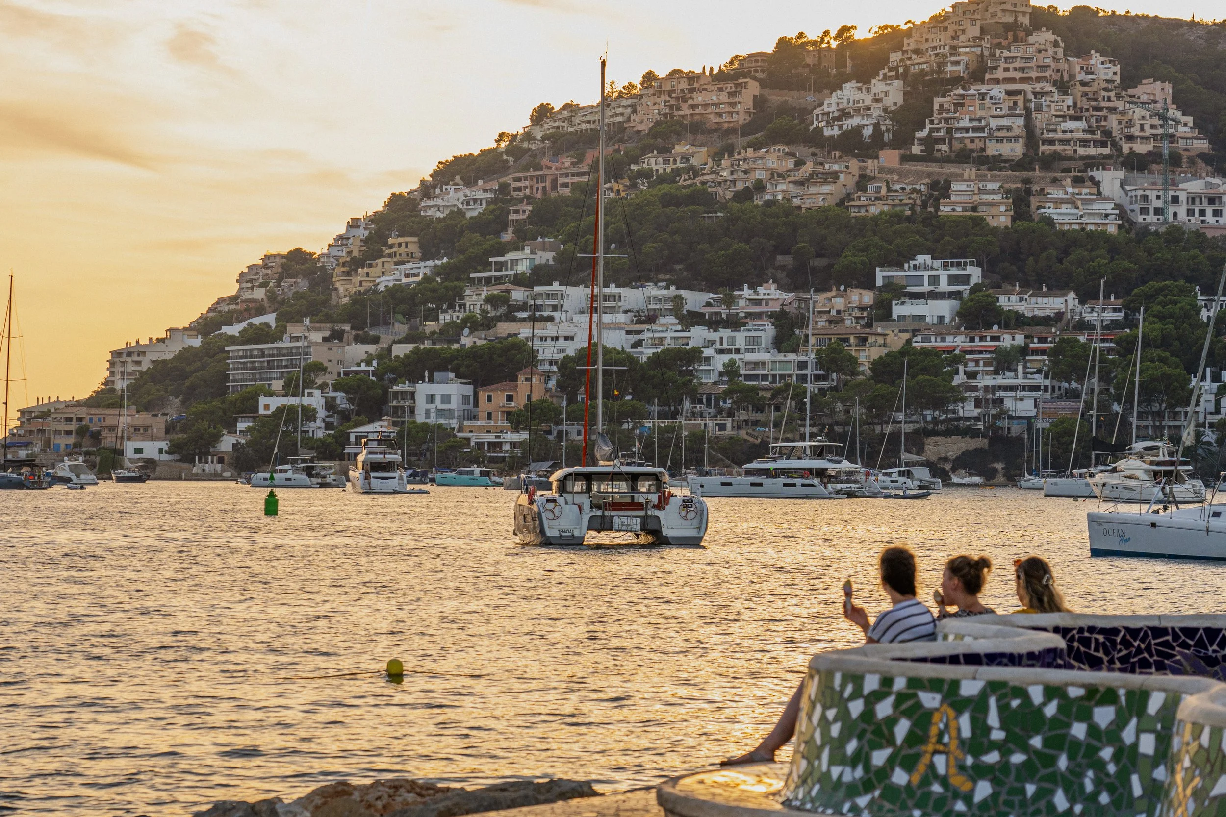 Vista de un puerto con yates y barcos, con una colina al fondo repleta de casas y un atardecer en el cielo