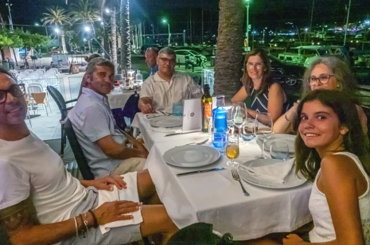 Grupo de siete personas sentadas en una mesa de restaurante al aire libre en la noche, con platos, vasos y bebidas en la mesa, ambiente iluminado con luces y palmeras al fondo.