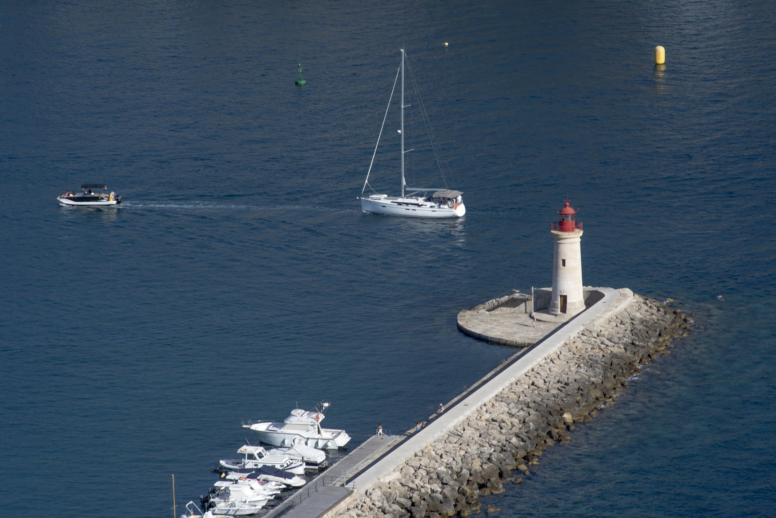 Un muelle con varios botes y un faro en la orilla junto a rocas en el mar azul