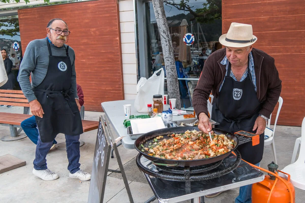 Dos hombres cocinando en exteriores, uno mezclando comida en un sartén grande y el otro observando, ambos llevan delantales y uno usa sombrero.