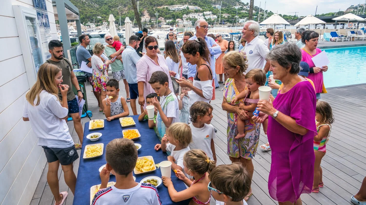 Una reunión en un muelle junto a una piscina, con niños y adultos, algunos con platos de comida y otros en línea, en un entorno de puerto y barcos.