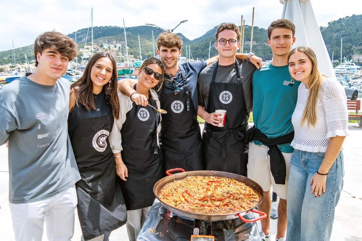 Grupo de amigos sonriendo con mariscos en un puerto, algunos con delantales, prepararán una paella.