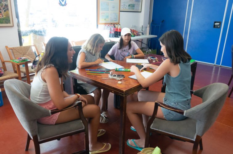 Cuatro niñas jugando y dibujando alrededor de una mesa en una sala con armarios azules y ventanas grandes.