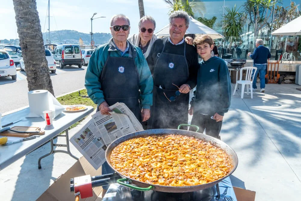 Cuatro personas, tres hombres mayores y un joven, levantan sus manos en celebración junto a una gran paella en una mesa al aire libre con palmeras y restaurantes, mientras que en el fondo se ven autos estacionados.
