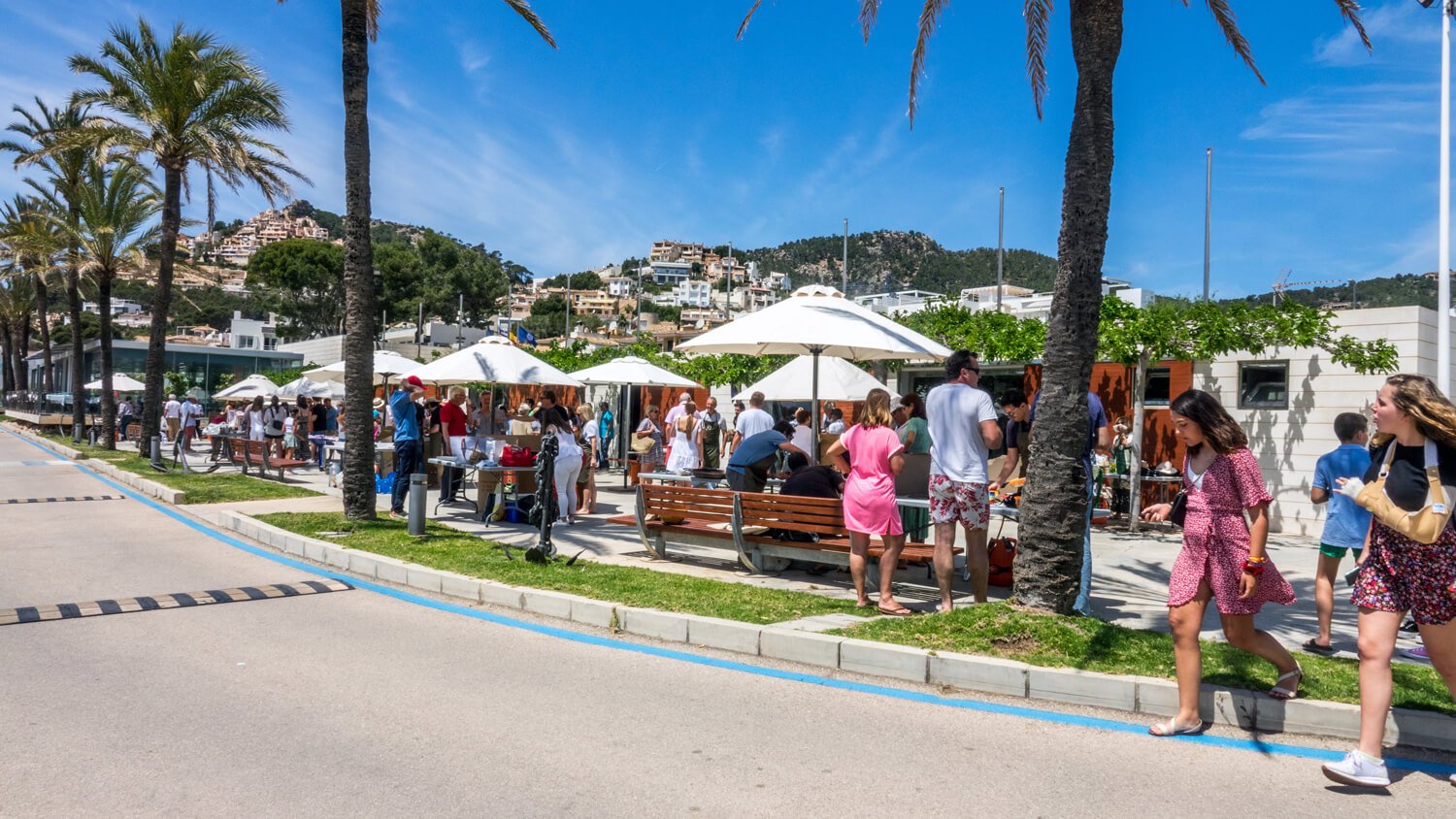 Personas en un mercado al aire libre con sombrillas blancas, árboles de palmeras, y colinas con casas en el fondo. Baja mañana soleada con cielo azul.