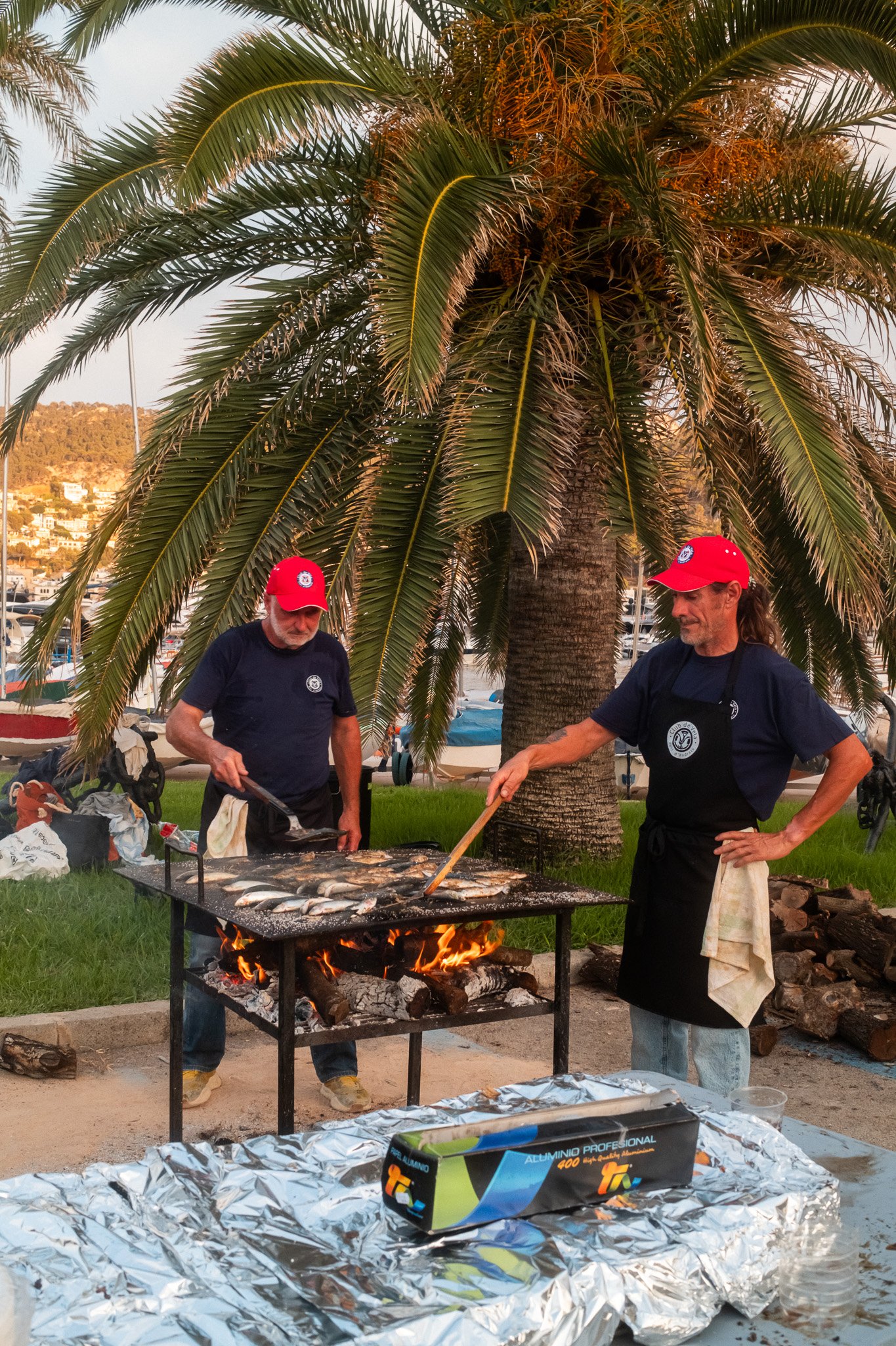 Dos hombres cocinando mariscos en una parrilla al aire libre bajo una palmera, con una ciudad y montañas en el fondo.