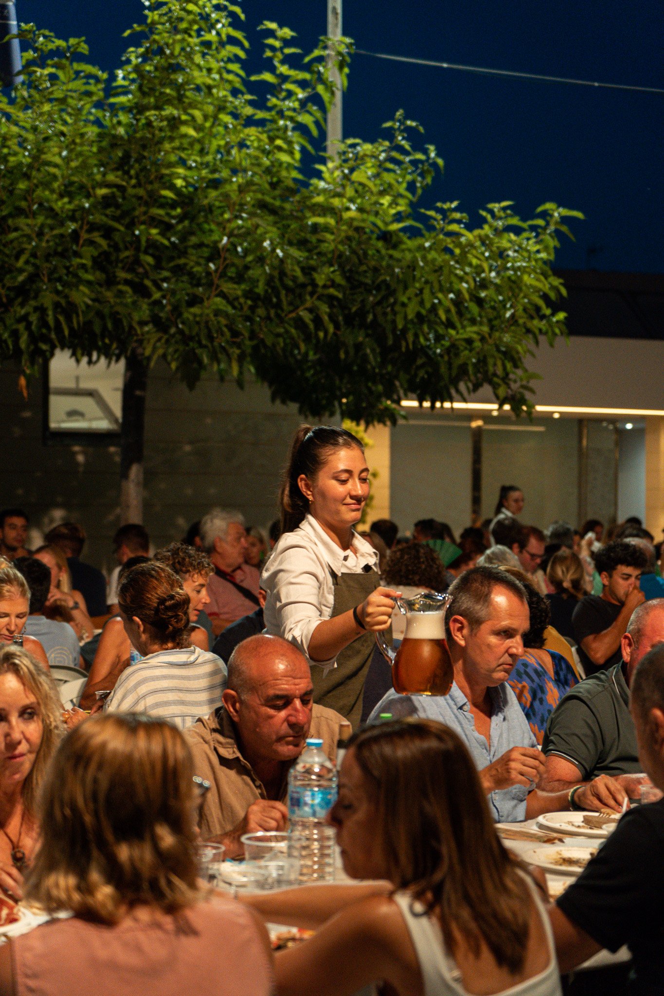 Una escena en un restaurante o evento social nocturno con muchas personas sentadas en mesas, sirviente sirviendo una jarra de cerveza a una mujer, ambiente iluminado, árboles y estructura moderna en el fondo.