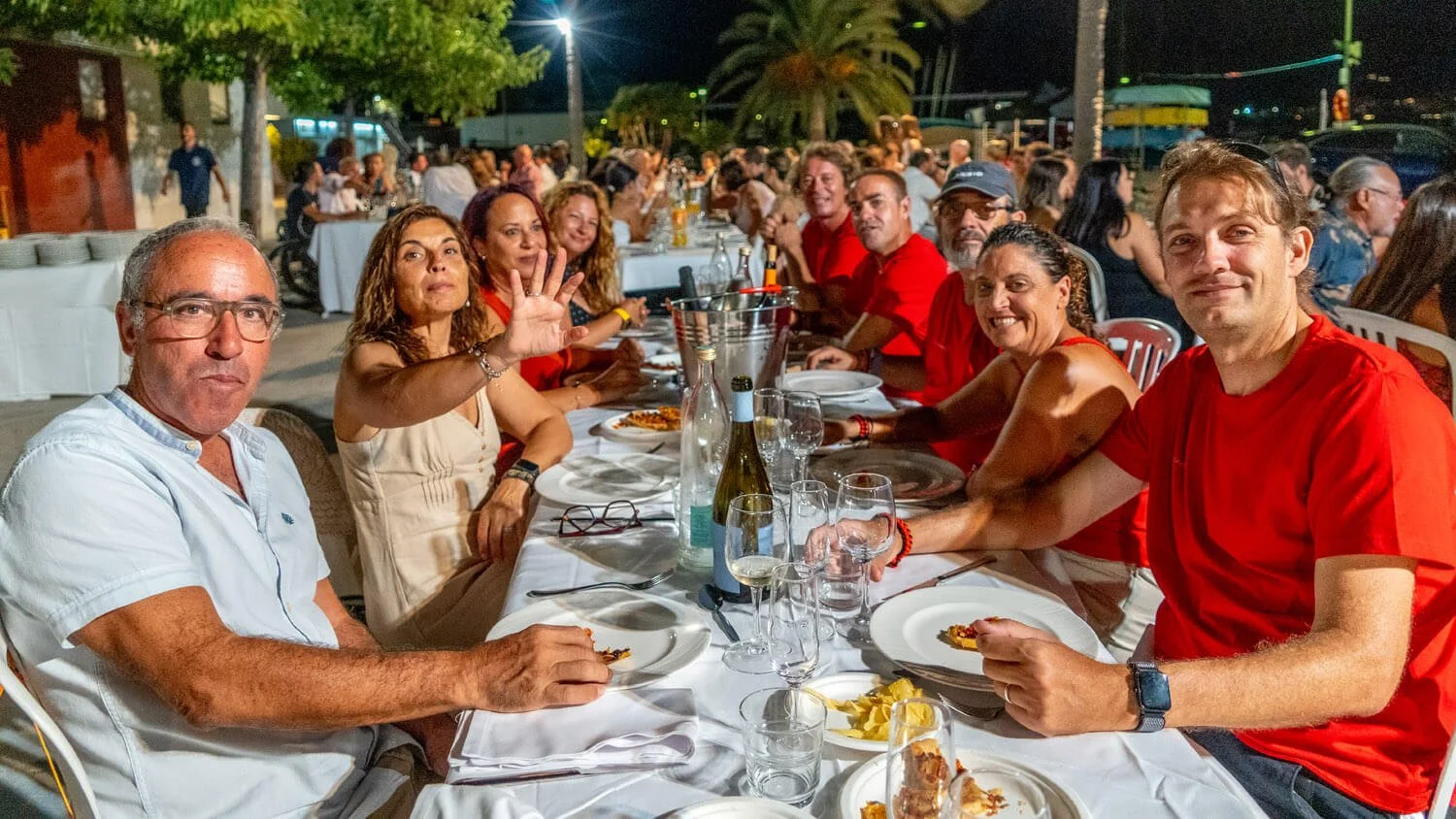 Grupo de personas en una reunión o cena al aire libre por la noche, sentados en una mesa con comida y bebida.