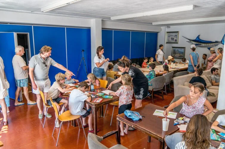 Niños y adultos en una sala de actividades, algunos sentados en mesas con libros y otros de pie, en un ambiente con decoración marina y una ballena en la pared.