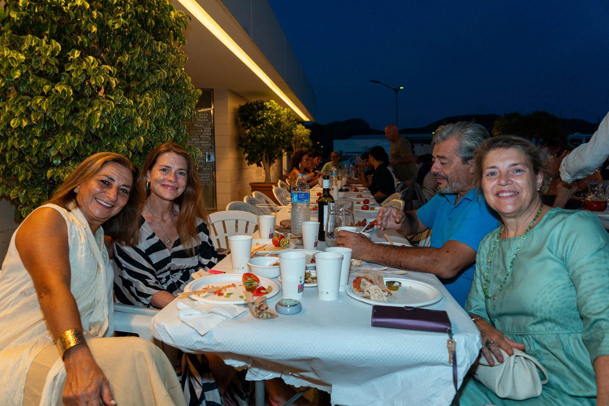 Personas sentadas en una mesa durante una comida al aire libre en la noche, con comida, bebidas y comida en platos desechables.