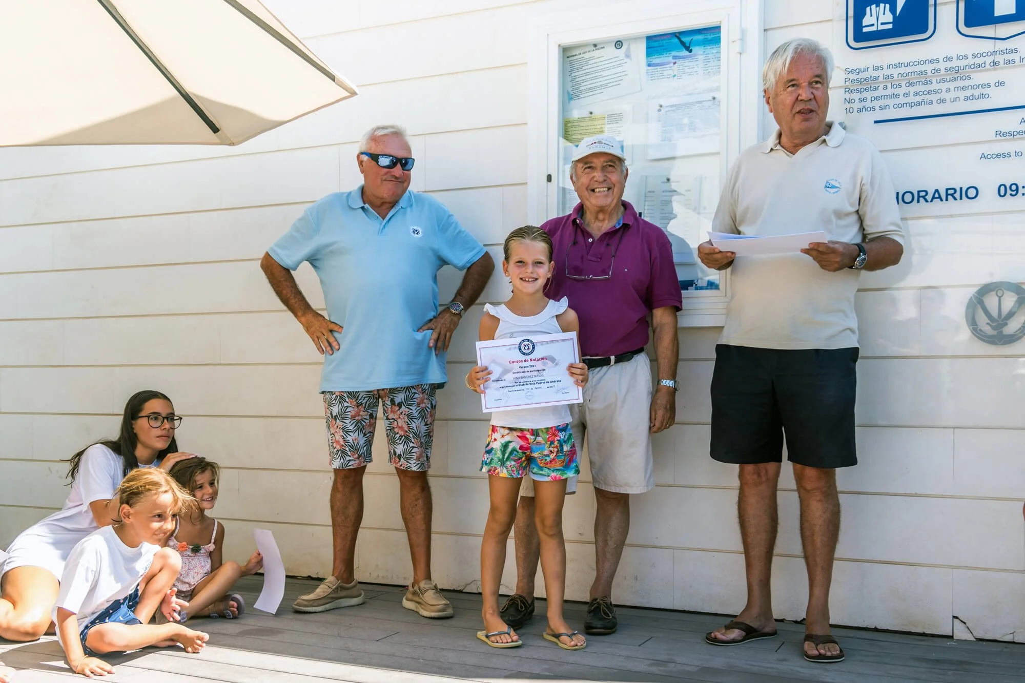 Grupo de personas, incluyendo adultos y niños, en una ceremonia de premiación al aire libre, con niños sentados en el piso y adultos de pie. Una niña sostiene un certificado, todos parecen felices y sonrientes.