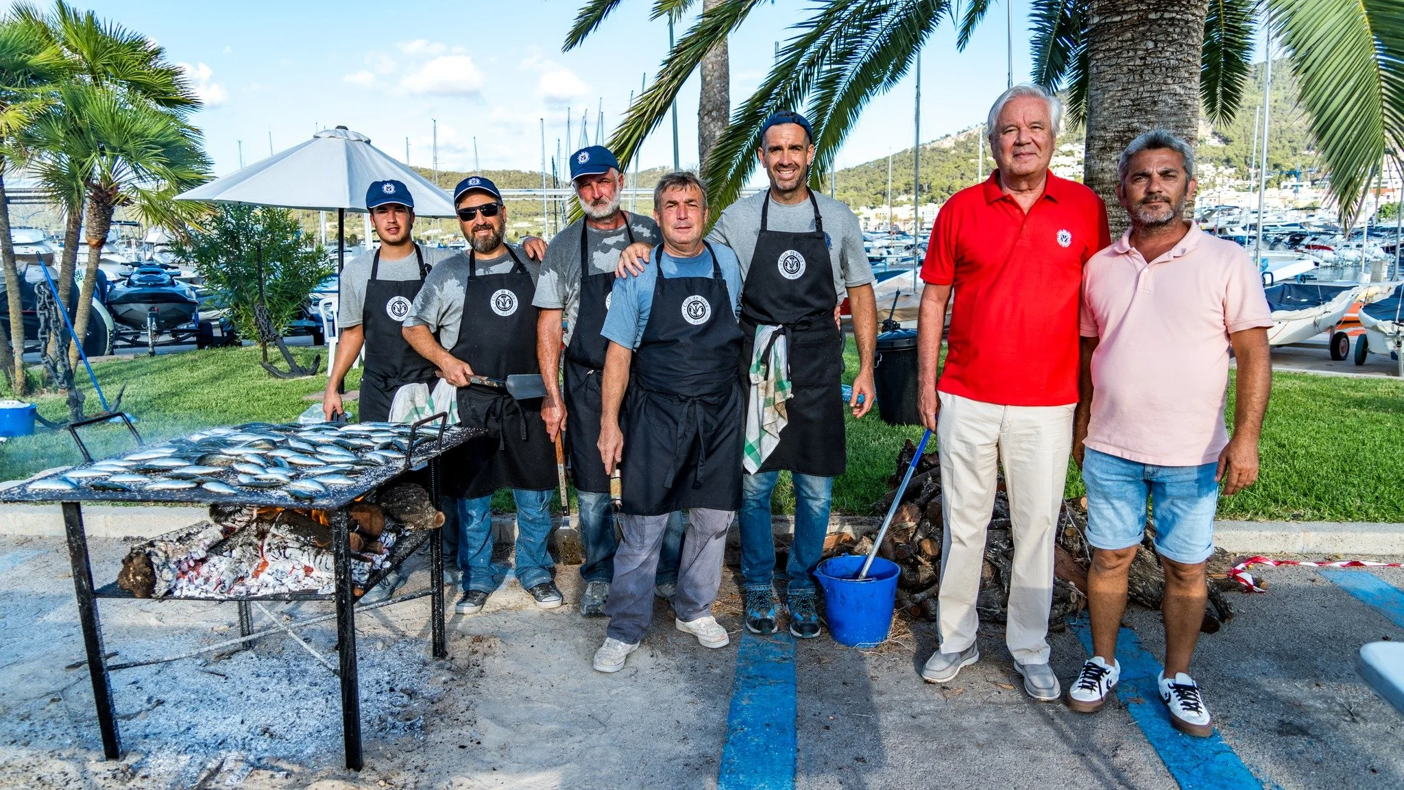 Grupo de personas, cocineros y amigos, en un muelle con palmeras, disfrutando de un asado de pescado, cerca de yates en un puerto.