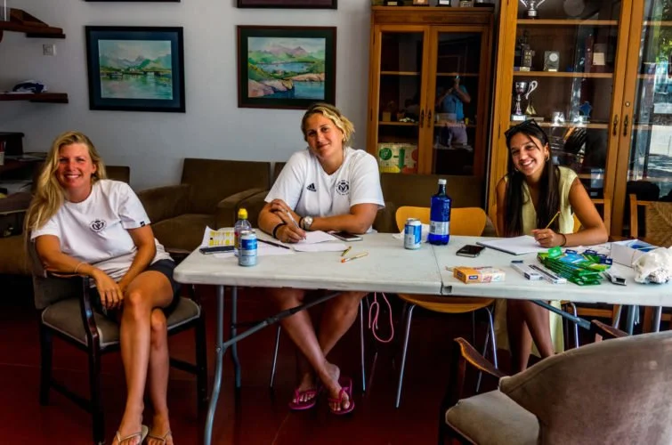 Tres jóvenes sentadas en una mesa en una habitación decorada con cuadros y muebles de madera, parecen estar en una reunión o estudio.