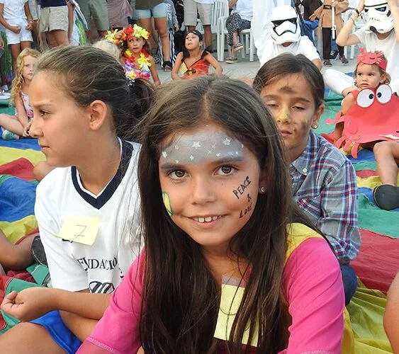 Niña con maquillaje de estrellas en la cara, sonriendo en una celebración con otras niñas disfrazadas y personajes de caricaturas en el fondo.