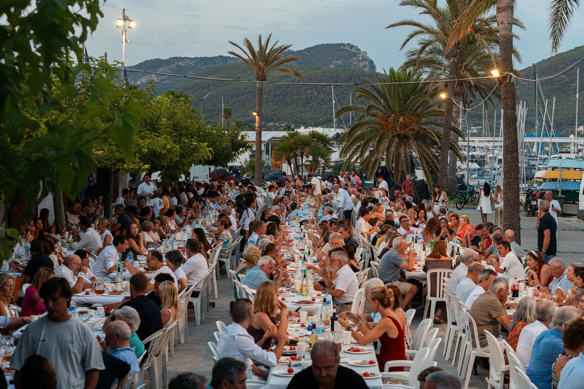 Una gran reunión al aire libre con muchas mesas y sillas, donde varias personas están comiendo y socializando en un muelle con yates y una montaña en el fondo, árboles de palmera y luces.