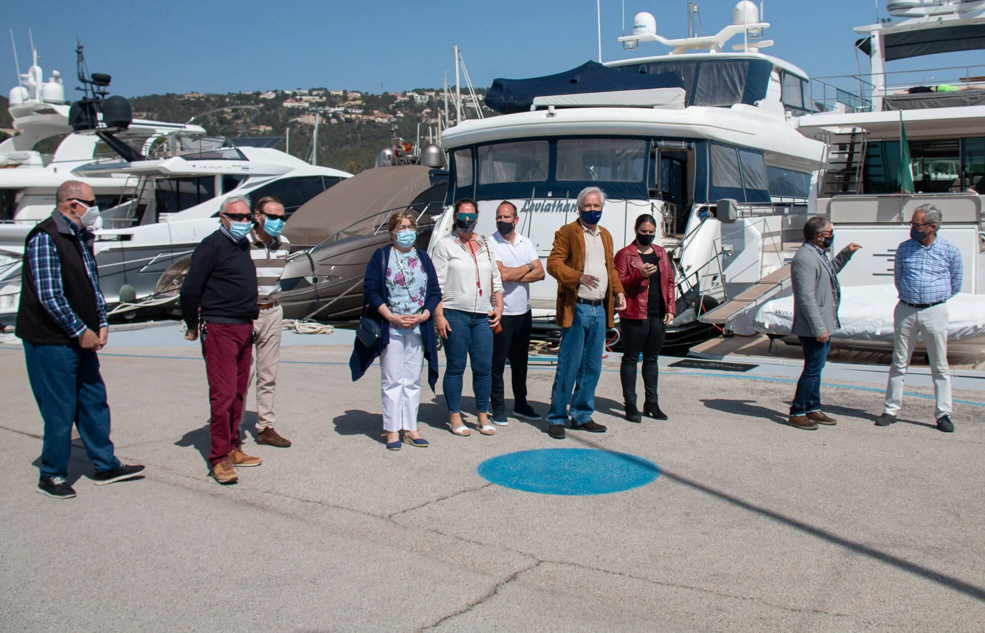 Grupo de personas con mascarillas en el muelle junto a yates de lujo, en un día soleado con colinas y casas al fondo.
