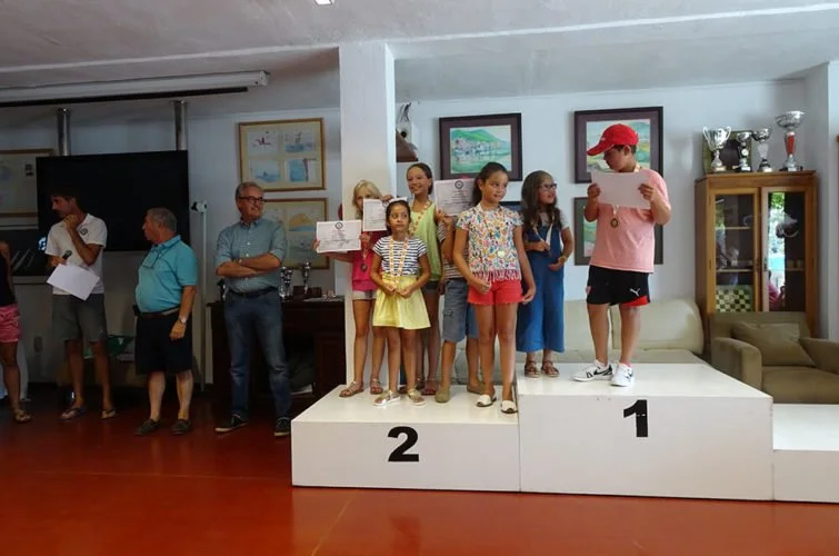 Niños y adultos en una ceremonia de premiación, con niños en un podio de primer y segundo lugar, leyendo certificados, en un salón decorado con cuadros en la pared.